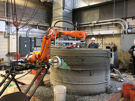 Four people look at an orange robotic arm as it moves above a cylinder concrete structure. 