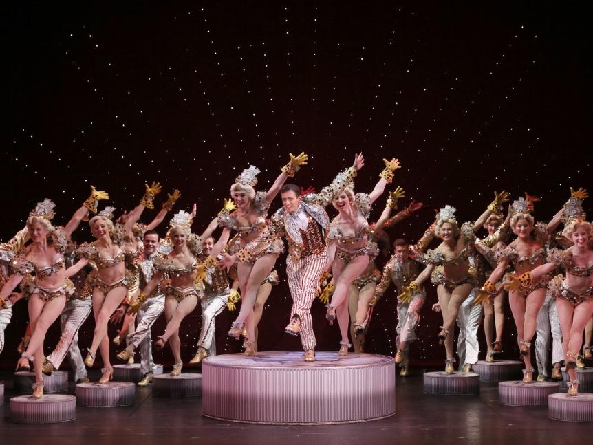 The ensemble cast for “42nd Street,” dressed in glitzy costumes, dance with their left legs extended in front of them and arms in up-and-down positions.