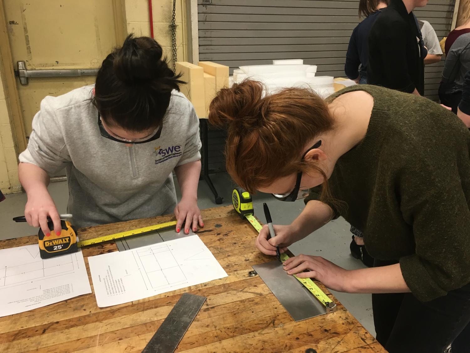 Two women measure sheet metal and mark the metal 