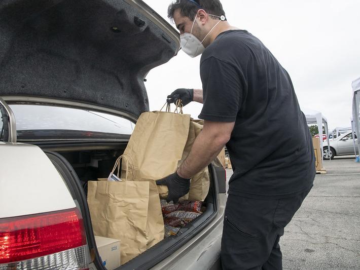 Person with face mask and rubber gloves loading groceries into back of car.
