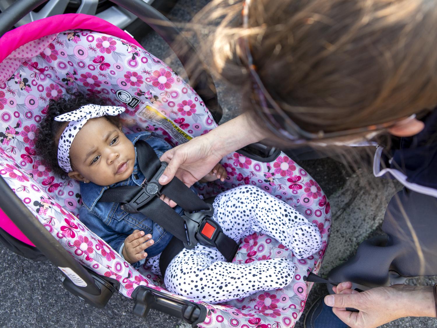 A woman leans over to check the car seat harness of a young child.