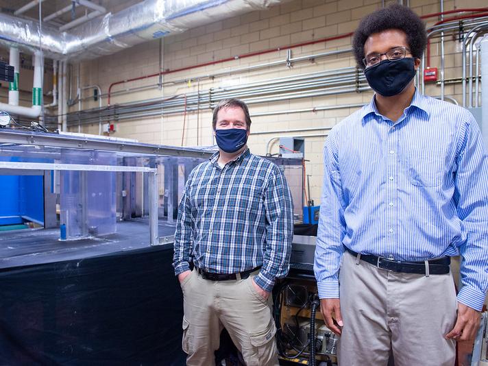 Two professors wearing masks stand besides an experimental wind tunnel in a laboratory.