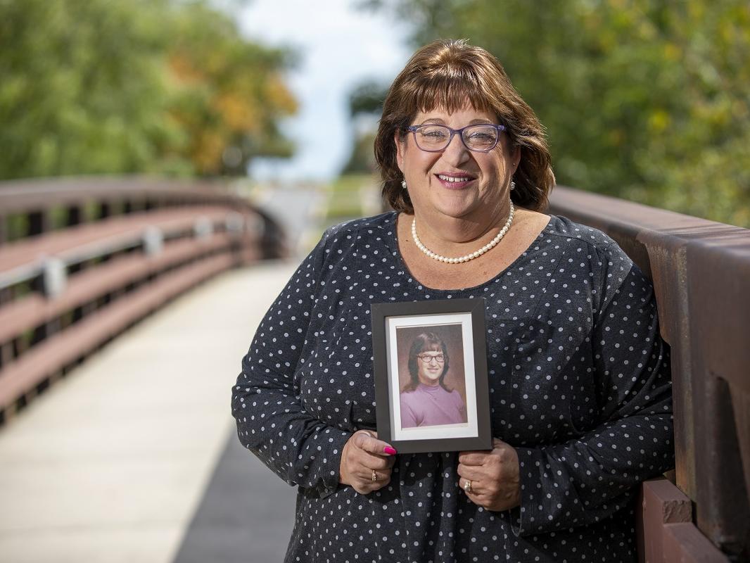 Carolyn Powell, who wears glasses and is smiling, smiles and holds a picture of herself taken in her early days at Hershey Medical Center. She is wearing a polka-dotted blouse and a string of pearls and stands on a bridge with trees in the background.