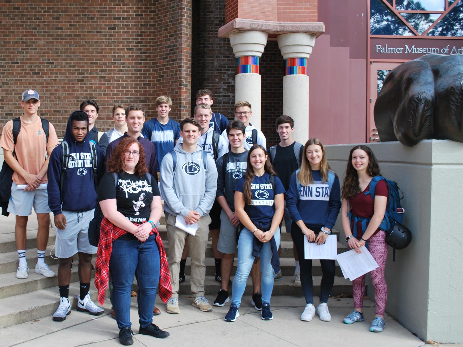 A group of students with their teacher stand together and smile on a sunny day in front of a brick building.