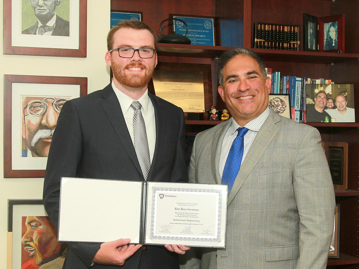 Two men stand in front of a book case. The man on the left holds a certificate. They are both smiling. 