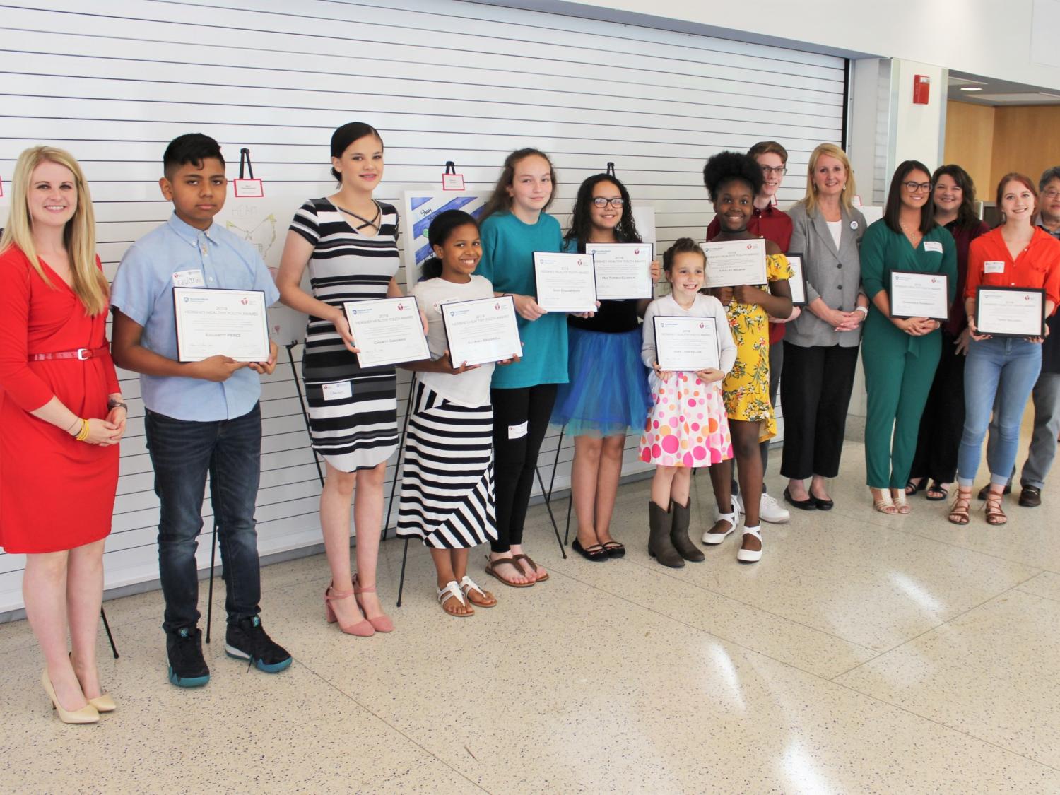 Fourteen people – 10 children and 4 adults – stand in a row to pose for a photo, all of them smiling. Each child holds an award certificate.