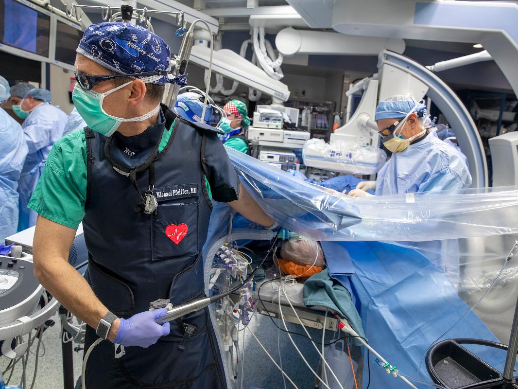 In the foreground, a surgeon watches a monitor while a second surgeon, background, prepares to insert a cardiovascular device on a patient