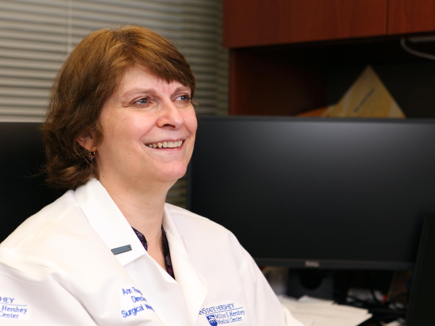 Dr. Ann Rogers is seated at a desk, wearing a white coat, and smiling.