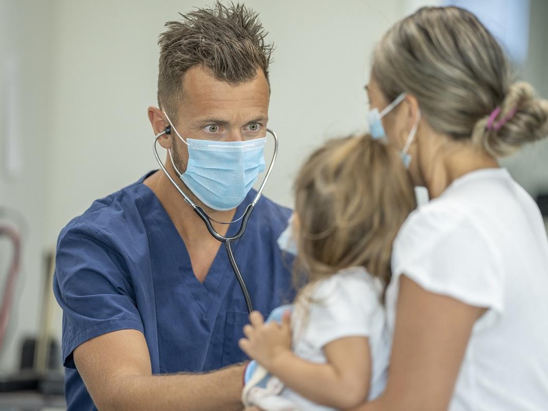 A nurse checks on a young patient's heartbeat. The little girl sits on her mother's lap while the nurse does the checkup.