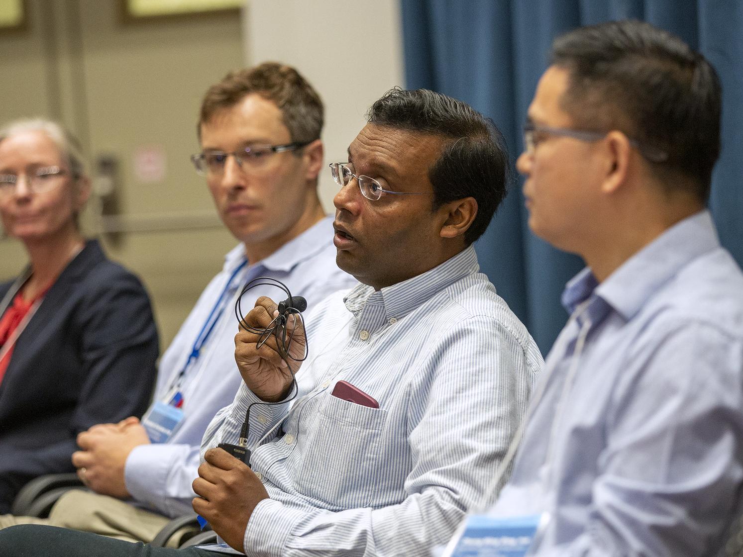 A row of five professional panelists is seated. The one in the center is holding a small microphone and speaking.