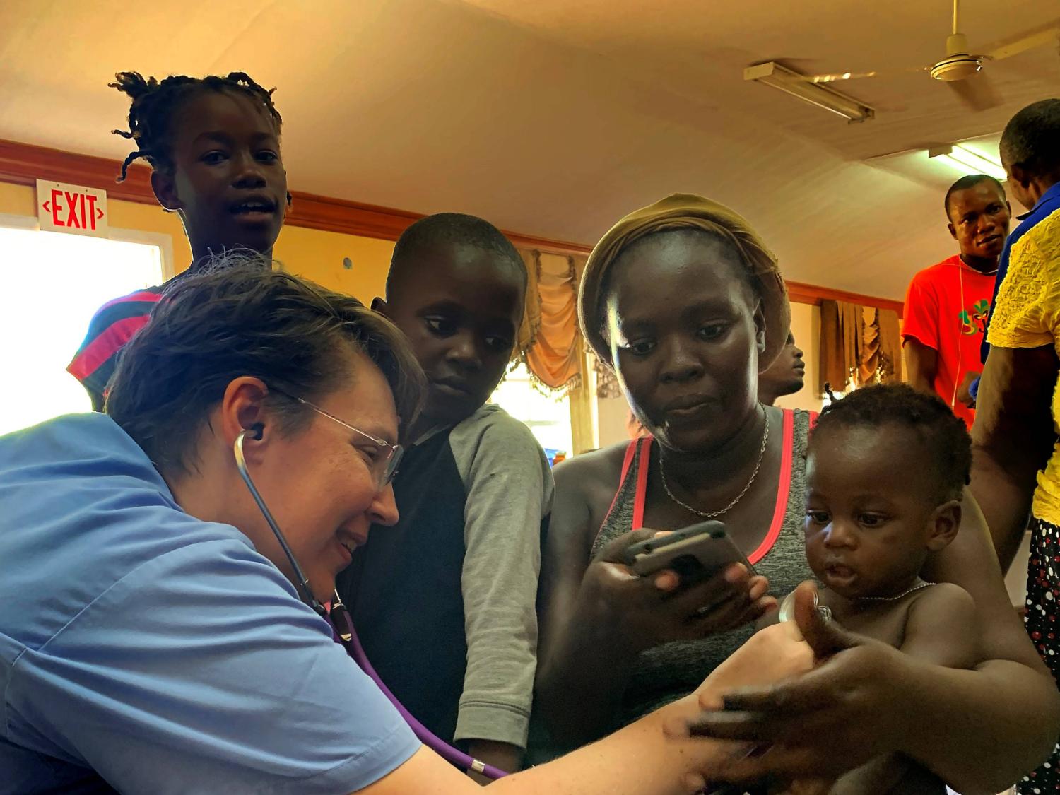 Alexandra Black, left, of Penn State Health St. Joseph, holds a stethoscope to the chest of a toddler who is sitting on his mother’s lap. She is wearing hospital scrubs and has short hair and glasses. Behind them two children watch.