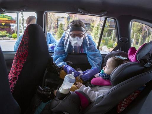 A woman leans into a car and touches the fingers of a young child. The woman is wearing a gown and face shield. The child is in a car seat and has a bottle in the cup holder next to her.