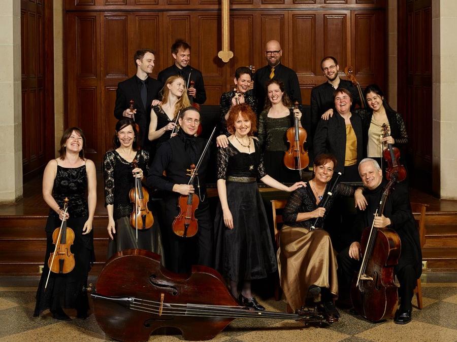 A group of musicians stand in a cluster while they hold their instruments and smile fot the camera.