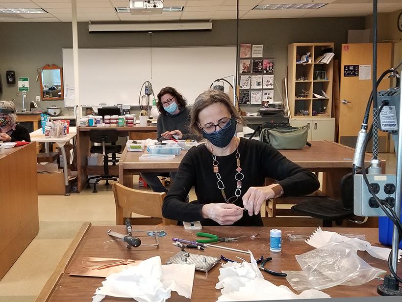 Three students wearing masks sitting at work tables and making jewelry.