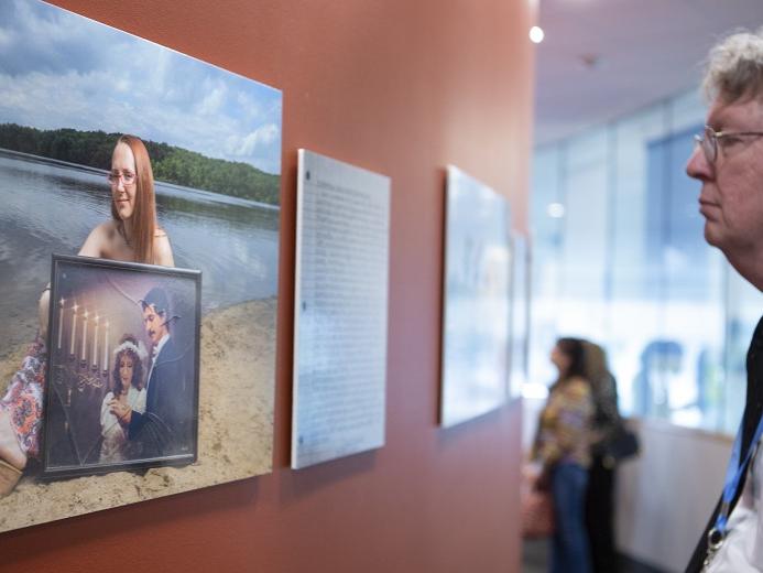 A man with glasses wearing a lanyard examines a photo of a bespectacled woman sitting on the beach of a lake lined with trees and mountains. The woman in the photo is holding another photo of man and a woman in wedding attire standing near a candelabra.