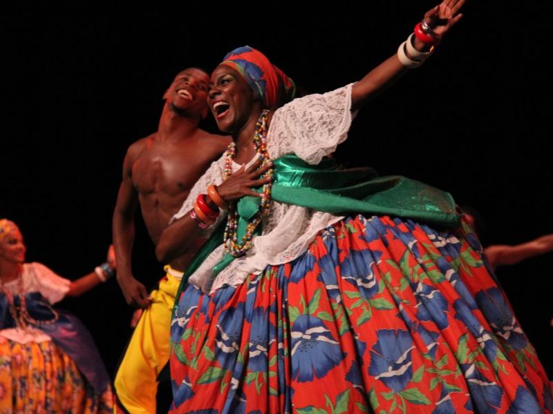 A dancer wearing a flower-printed full skirt and headwrap and colorful bangles and beads smiles and sings while she holds her right hand to her chest while outstretching her left hand.