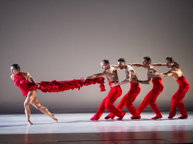 Four males dancers stand one behind the other while the first in line holds on to a fabric train worn by a woman dancer standing in front of them.