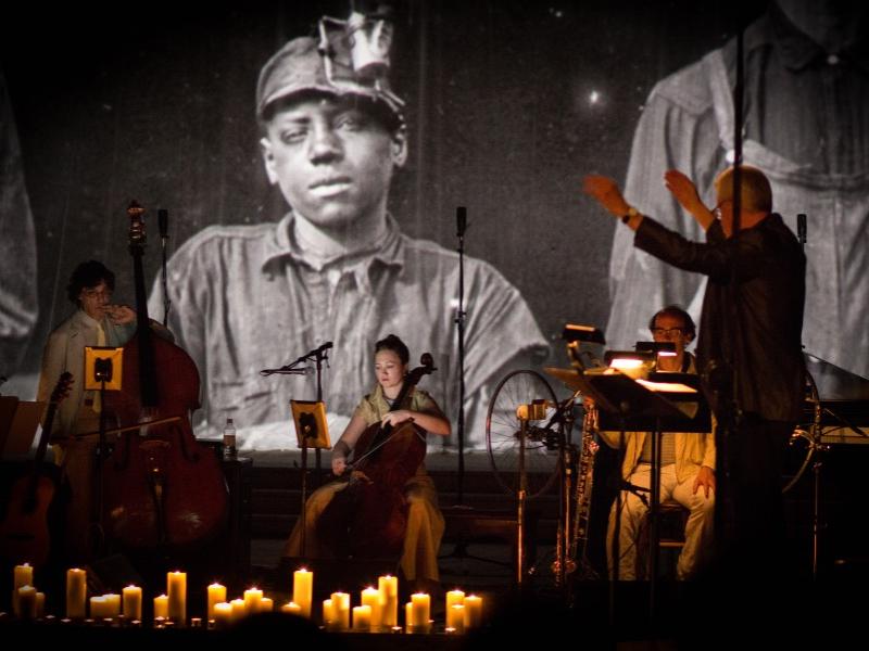 Musicians from Bang on a Can All-Stars sit on stage and perform while images of coal miners are displayed behind them.