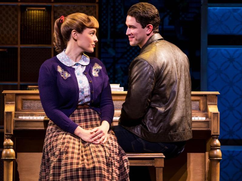 A woman and man sit on a piano bench and make flirtatious faces at each other.
