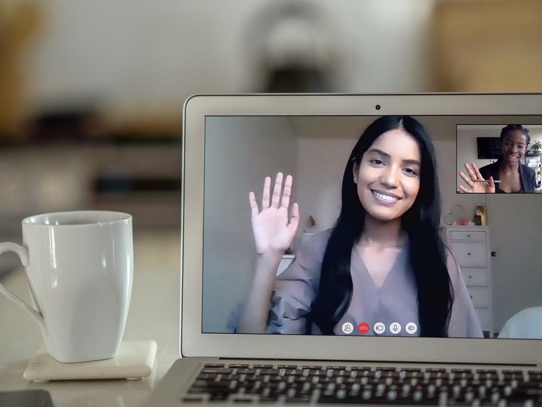 A woman in a video chat on a laptop screen waves to another, shown in a smaller window on the screen. A coffee cup is next to the laptop.