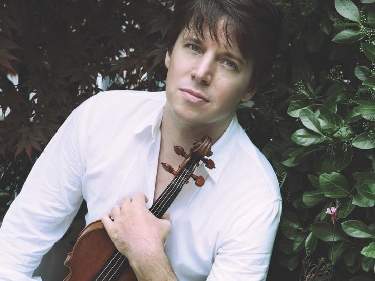 A musician sits on a chair and holds his violin facing the camera in his left hand.