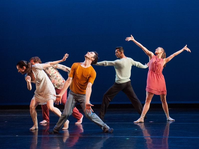 Dancers dressed in casual clothing and wearing only socks make various gestures with their outstretched arms.
