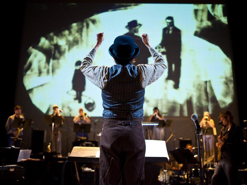 Darcy James Argue holds his arms up while conducting his big band, which stands with instruments in front of a hand-painted background.