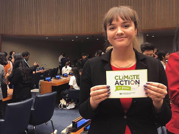 A smiling woman stands in a crowded room, with tables, chairs and other people visible in the background. She holds up a "Climate Action for Peace" sign.