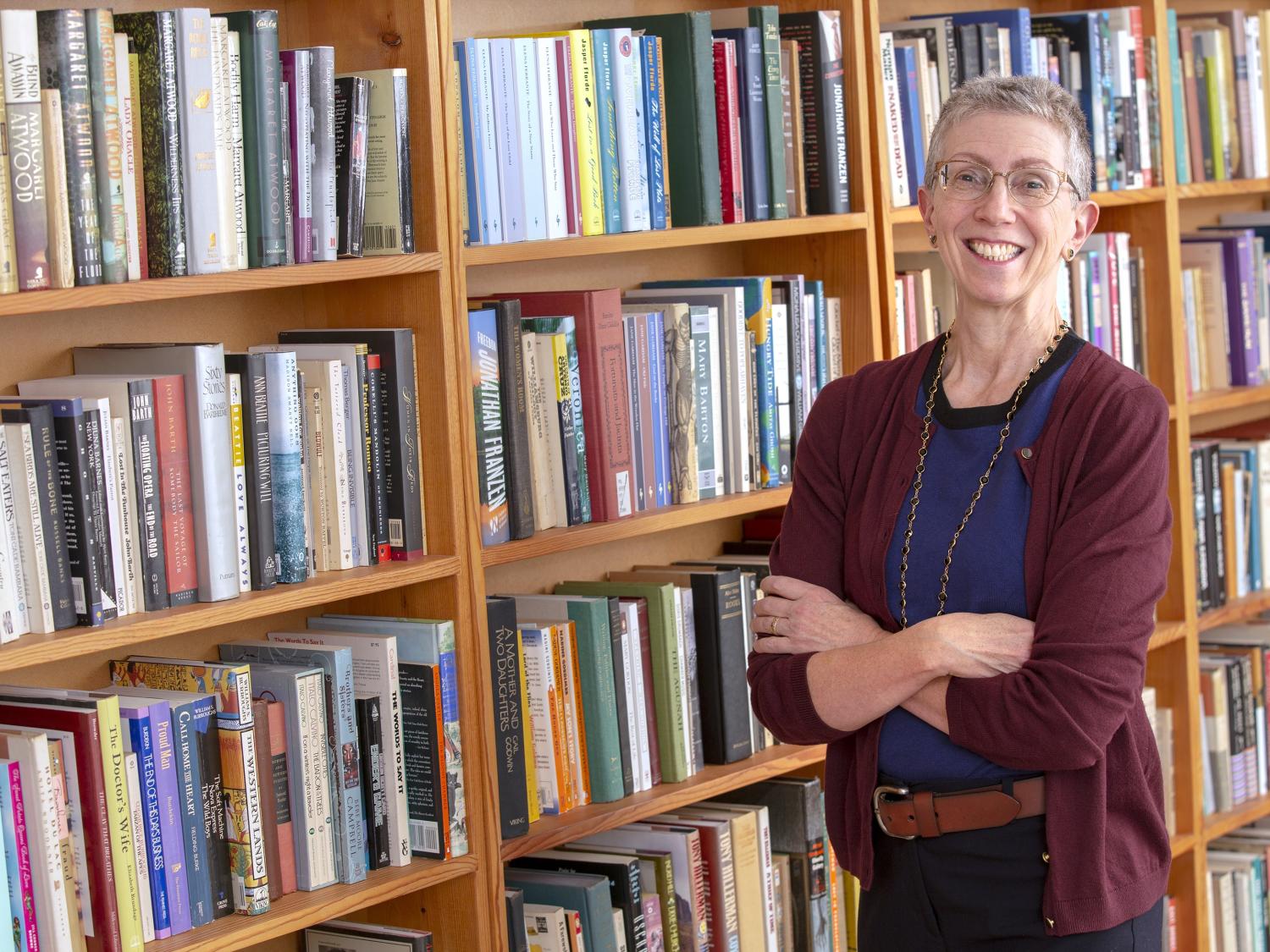 Bernice Hausman stands in a library with her arms crossed, smiling. She has short, gray hair and is wearing glasses, a sweater, a T-shirt and casual pants. Bookshelves filled with books are to her left.