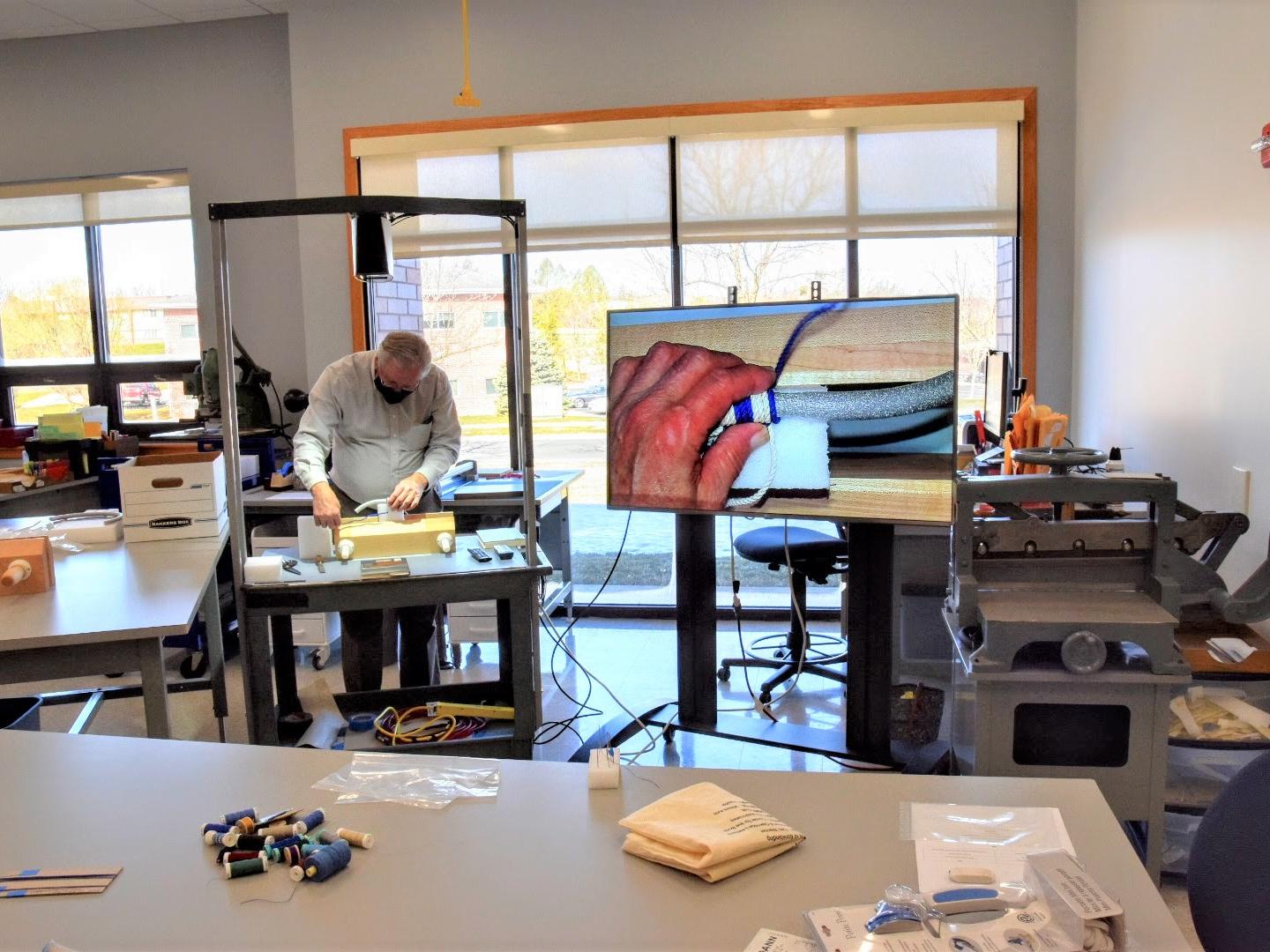man standing at small table in work room with hands shown close up via overhead camera video display demonstrating work with two alternating colors of thread around round finger-thickness length of foam