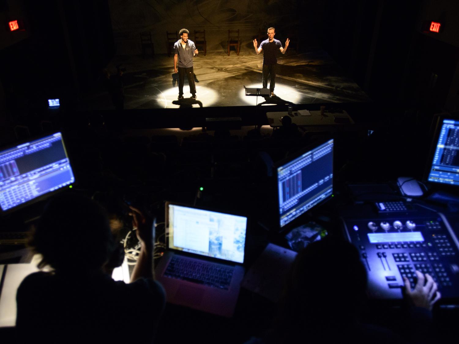 two male actors standing in spotlights onstage with production computer screens and lighting controls in foreground