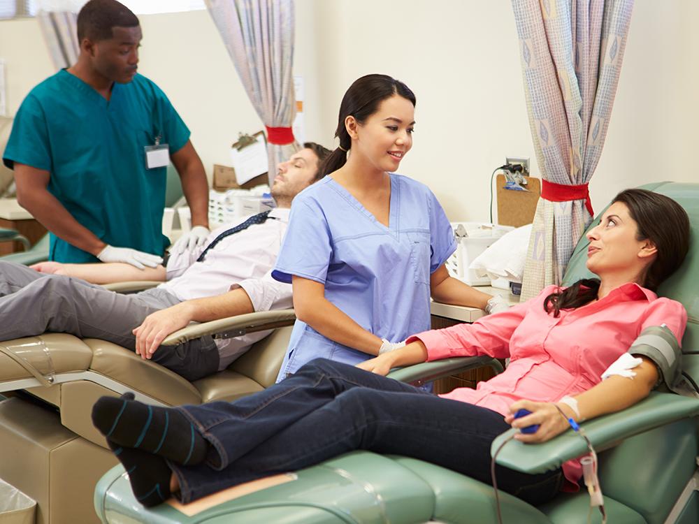 In the foreground, a female nurse tends to a female patient who’s donating blood. In the background, a male nurse tends to a male patient who’s donating blood.