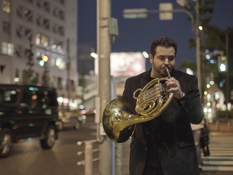 A french horn player holds his instrument while walking in the street.