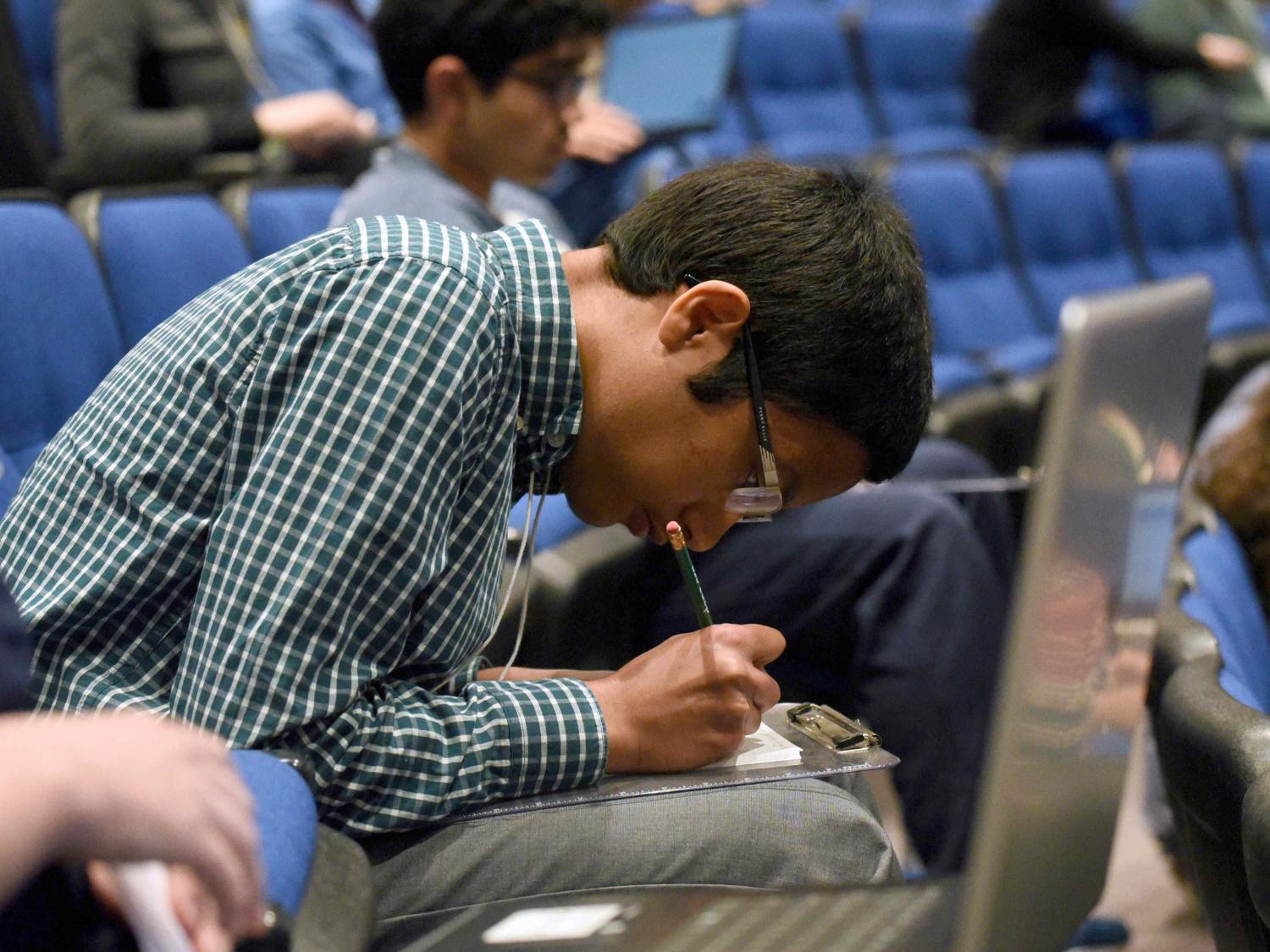A student bends over a clipboard on his lap and writes during the 2019 National Brain Bee, held April 12-14 at Penn State College of Medicine. He is seated in an auditorium, surrounded by other students. He wears a plaid shirt, gray pants and glasses.