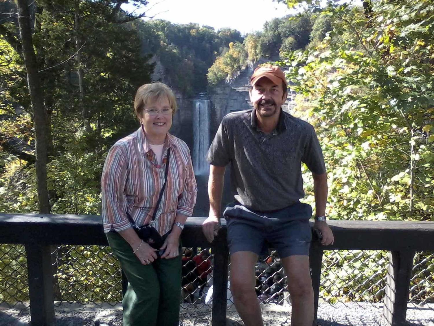 Glenna and J. David Brensinger lean against the side of a bridge. In the background are trees and, at a distance, a large waterfall.
