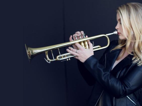 Bria Skonberg, shown in profile, holds her trumpet to her mouth and faces left.
