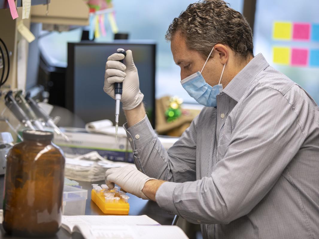 Virologist Nick Buchkovich, seated at a lab bench, uses pipetting equipment. Additional lab equipment and various books and papers are also on the bench.