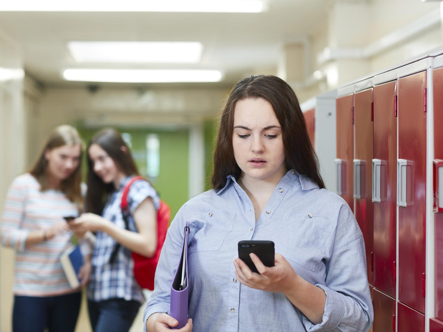 A girl looking at her phone as two teens giggle in the background