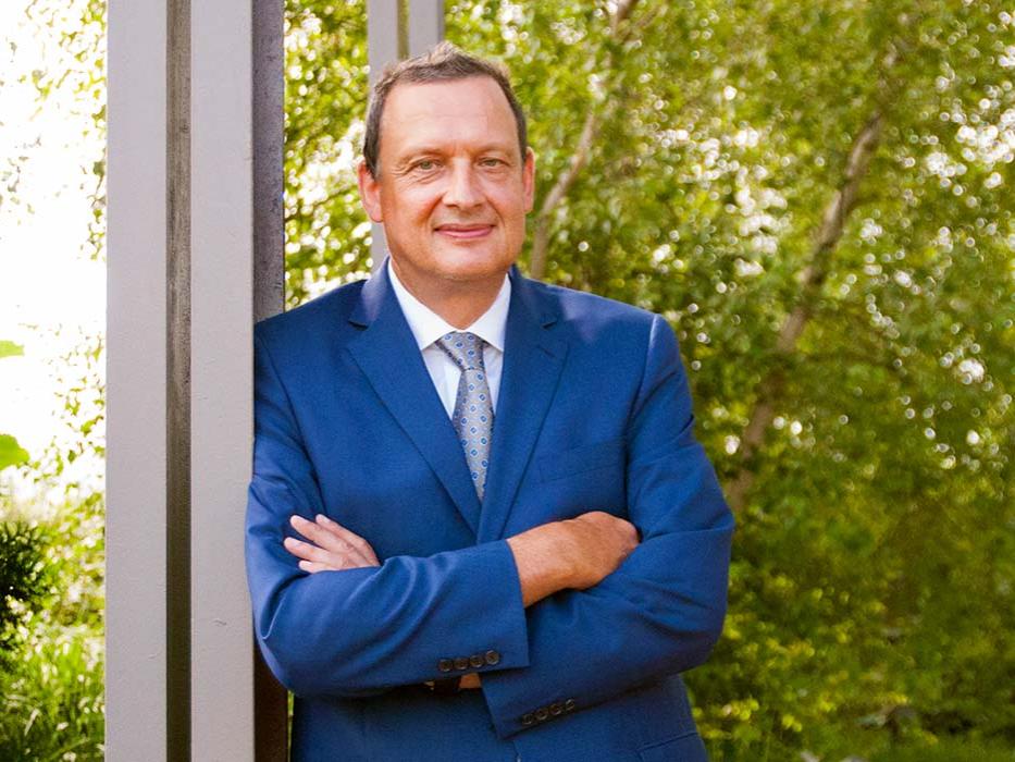 Michael Büsges in a blue suit with his arms crossed. Green foliage is in the background.