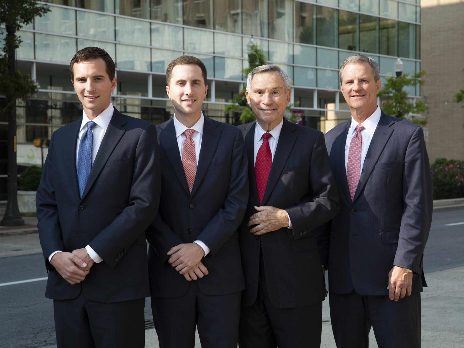 Four men in suits and ties pose for a photo in front of an office building.