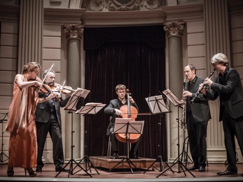 Three string and two wind musicians dressed in formal clothing stand in a semicircle on a stage and perform.