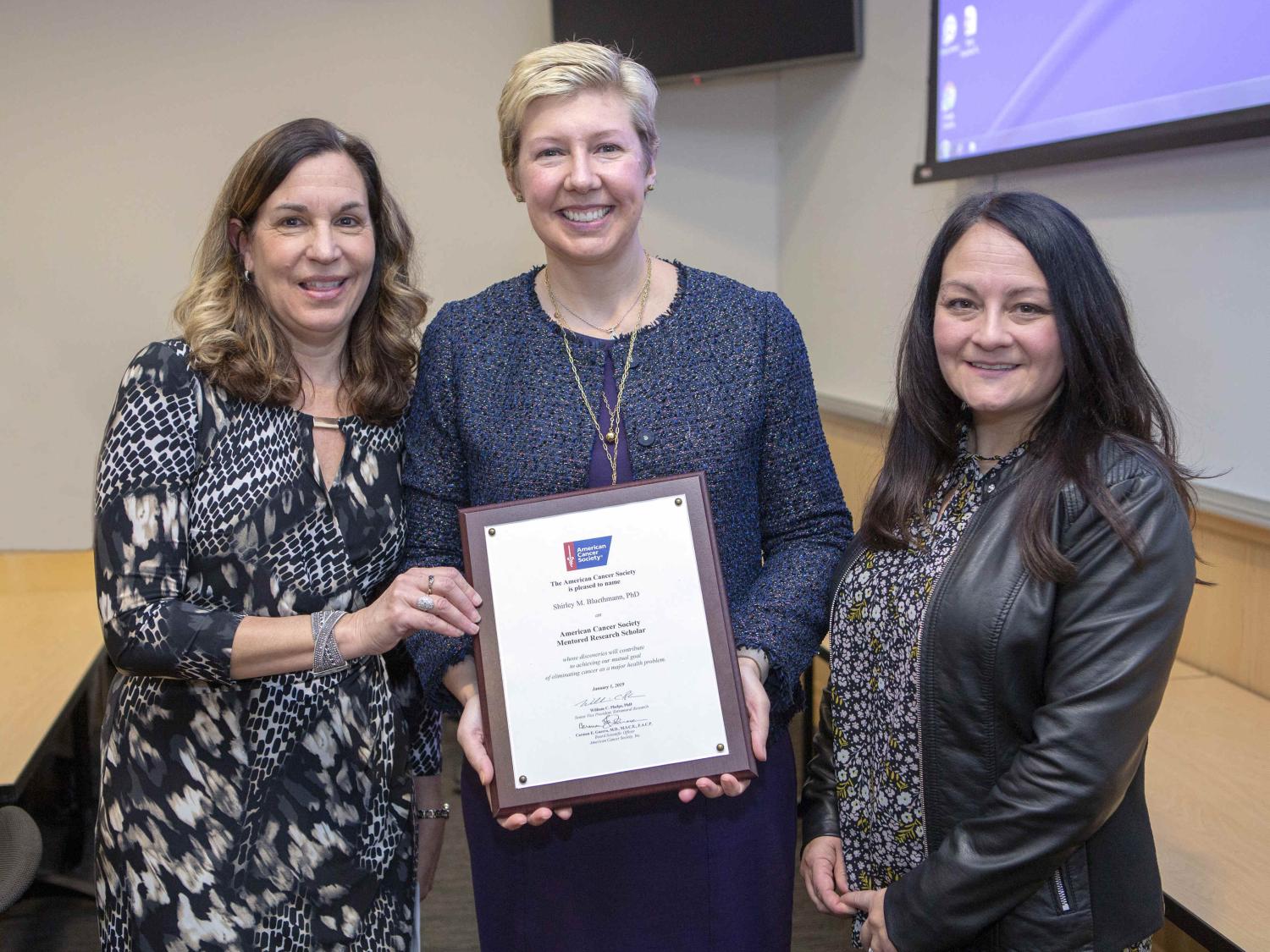 Three women in professional attire pose for a photo in a conference room. The woman in the middle holds a framed certificate bearing the American Cancer Society logo. A portion of a table and parts of two wall-mounted video monitors are in the background.