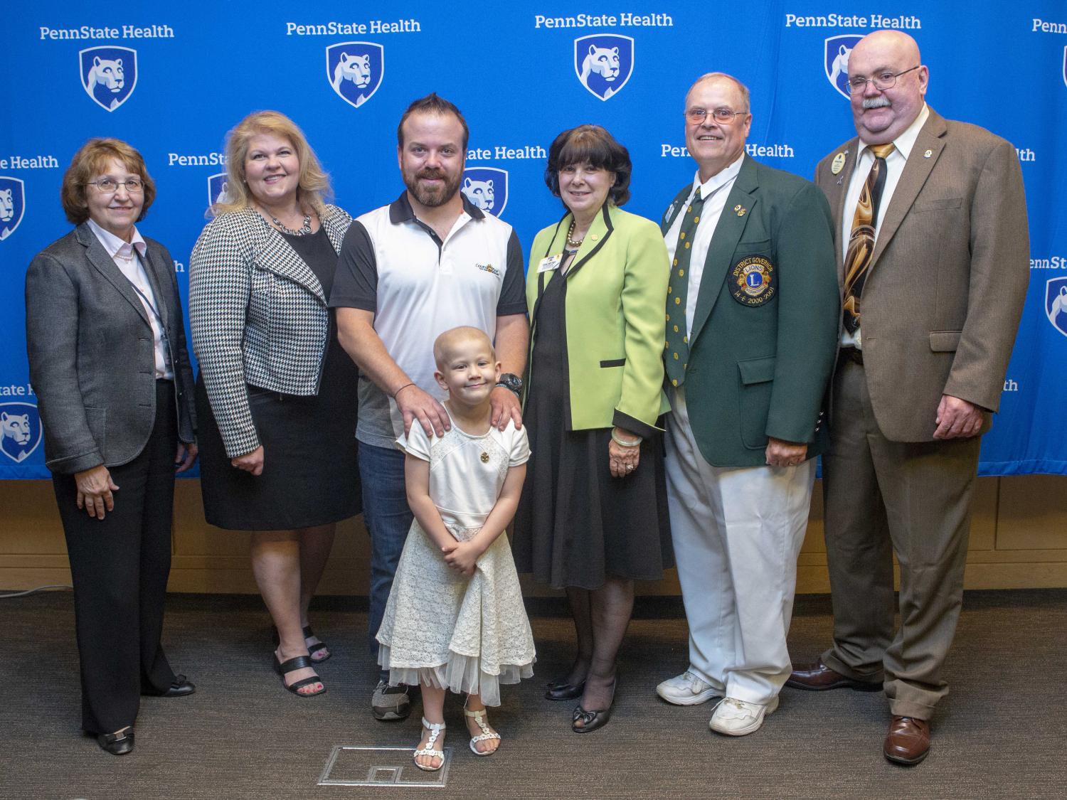 Six adults and one young girl pose for a photo. The girl is in front, smiling. The backdrop has several 'Penn State Health' logos.