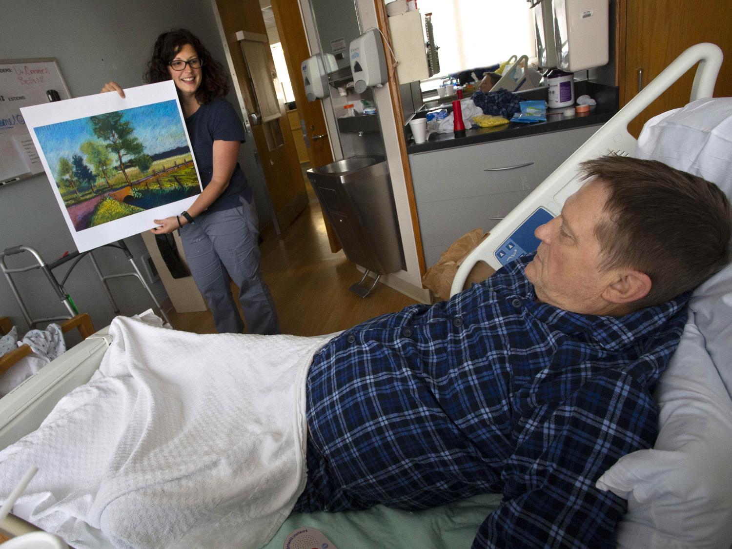A man lays in a hospital bed. A woman stands at the foot of the bed holding a painting, showing it to him.