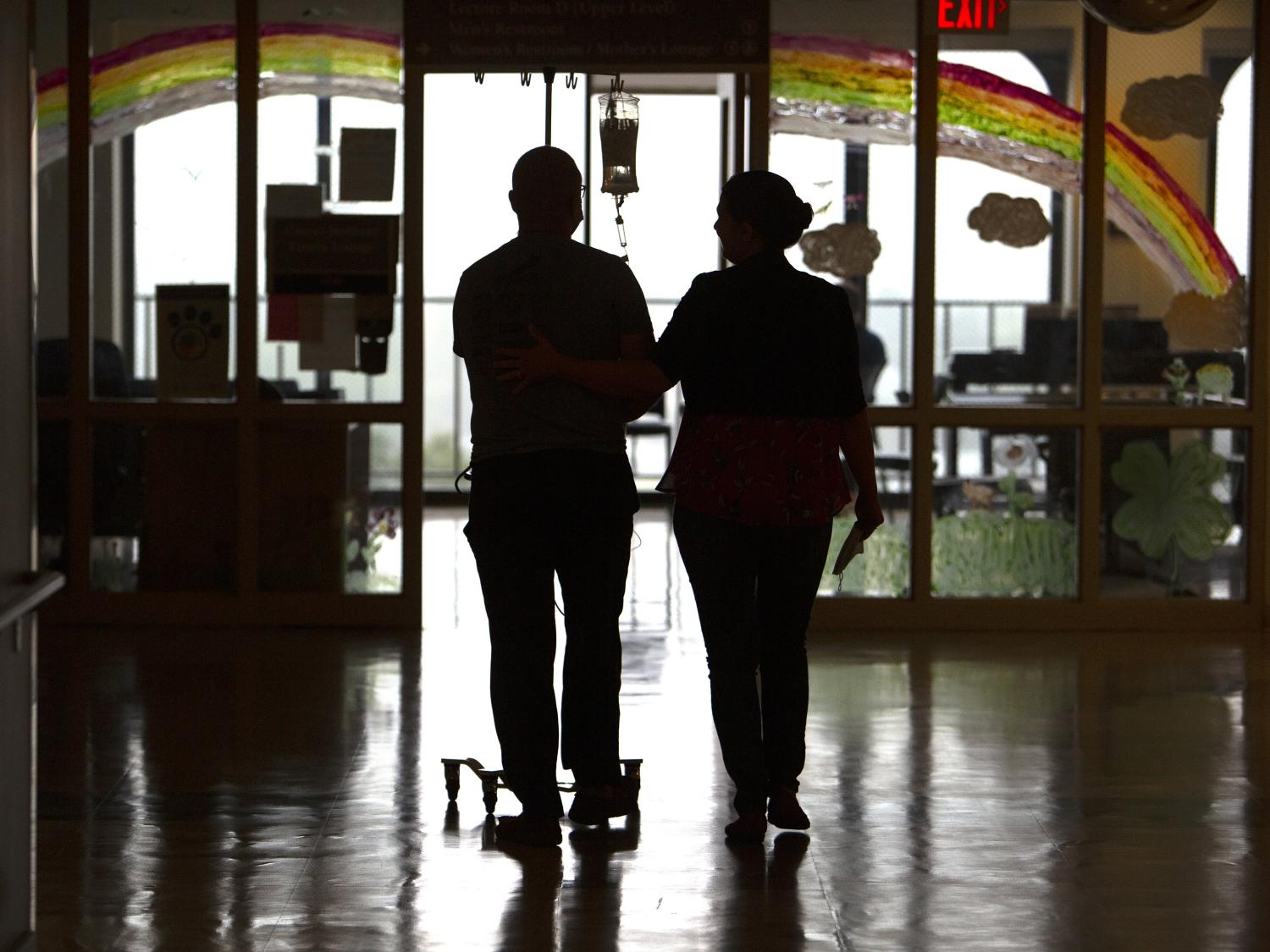 Tim Card and his wife, Tricia, walk the hallways of Penn State Cancer Institute after he receives CAR-T cell therapy infusion. Their backs are toward and the camera, and they are silhouetted against a window. Tricia puts her hand on his back. Tim is pushing an IV pole, and an IV bag with medication hangs from the pole. A rainbow is painted on the window.
