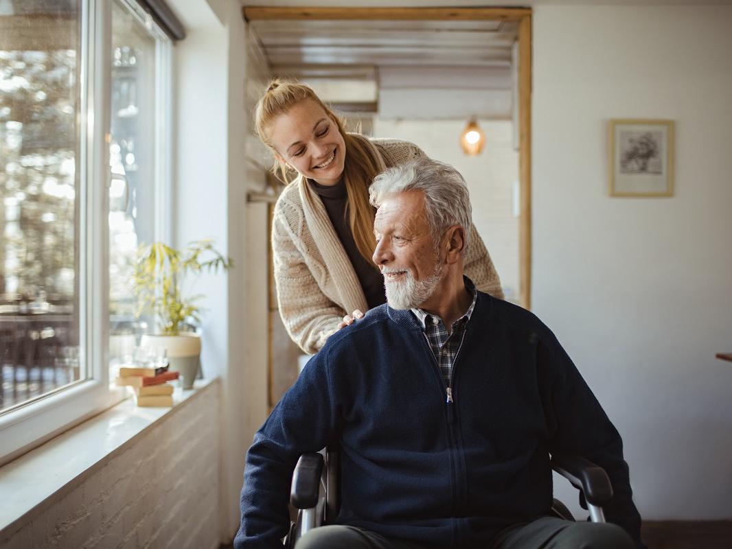 A woman pushes an older man in a wheelchair, as both smile. They are next to a sunny window.