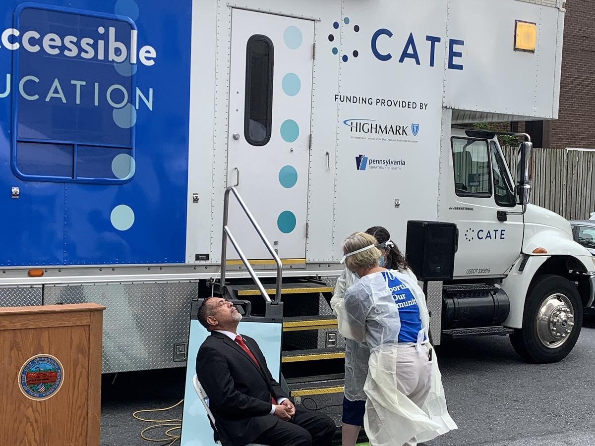 Eddie Moran leans back in a chair while two women in PPE stand in front of him. Behind them is a truck with the word CATE on the side.