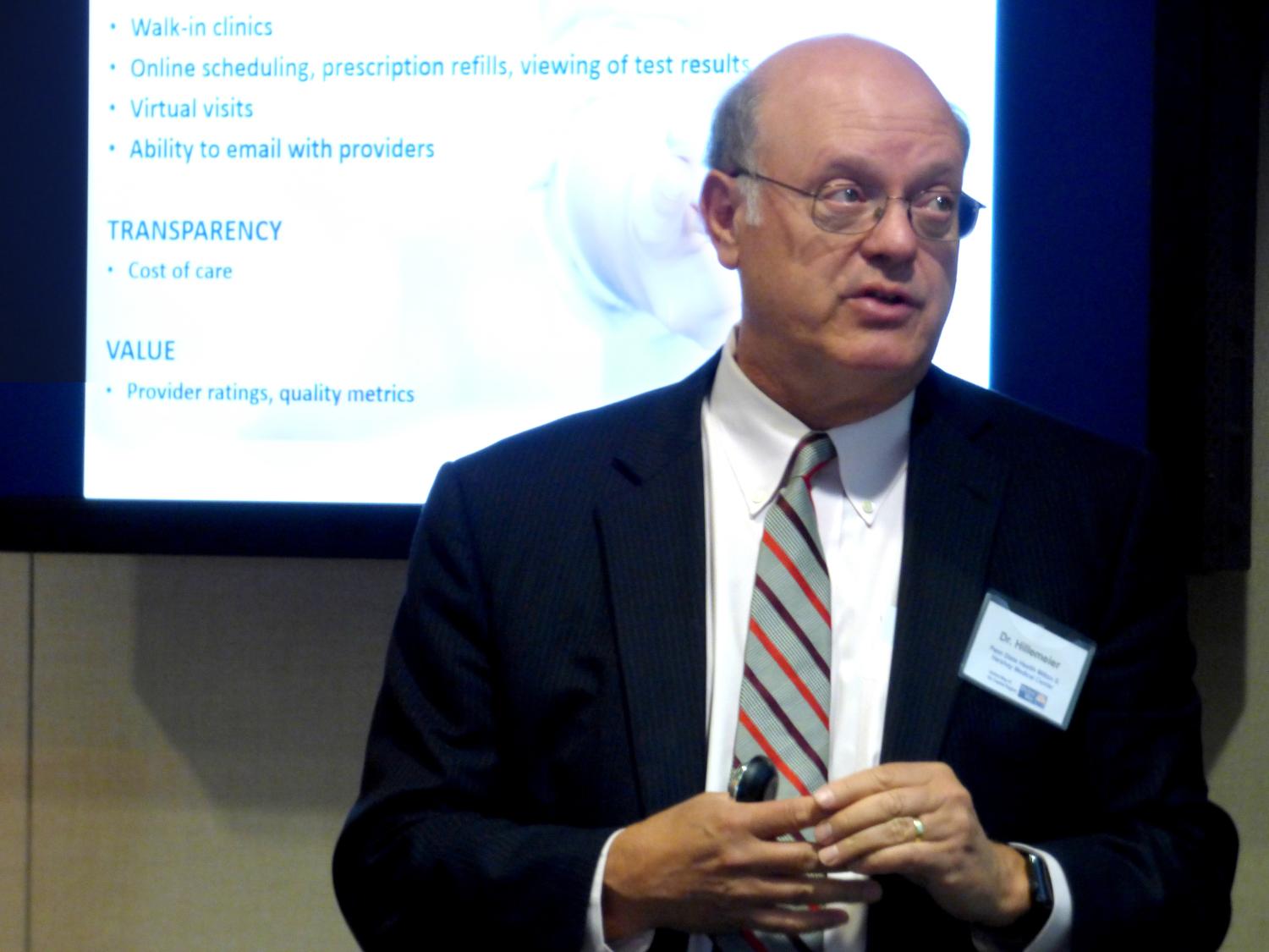 Dr. Craig Hillemeier wears a suit and stands in front of a screen bearing a presentation, speaking.