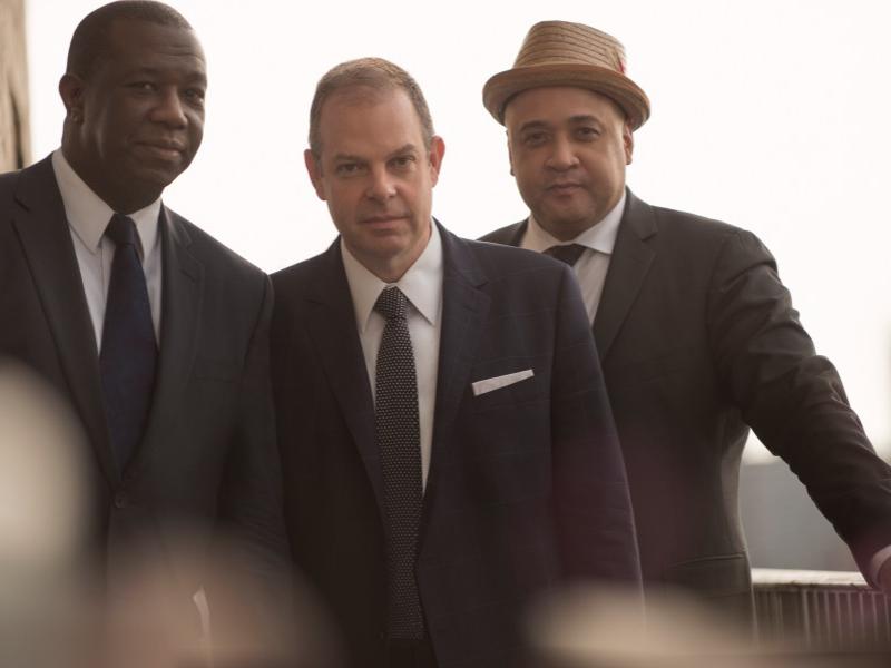 Three men in suits stand in front of a window.