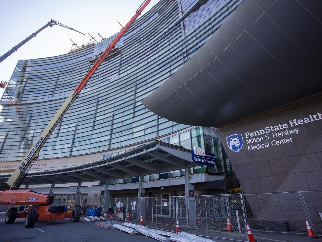 Two cranes sit in front of the entrance to the Children’s Hospital. A chain link fence wraps around the front of the building. Construction cones and other items are scattered about.